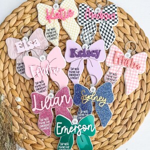 May include: A selection of bow-shaped keyrings in diverse colours and patterns, each bearing a name and the message: "Tap with phone for emergency contact." The keyrings showcase glitter, gingham, and checkered designs.