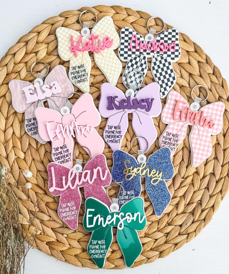 May include: A selection of bow-shaped keyrings in diverse colours and patterns, each bearing a name and the message: "Tap with phone for emergency contact." The keyrings showcase glitter, gingham, and checkered designs.