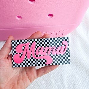 Pu&ograve; includere: Un biglietto a scacchi rosa e nero con la parola "Mama" in lettere corsive rosa.