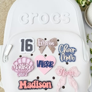 May include: White Crocs backpack adorned with decorative charms. Charms include a number 16, glittery bows, a seashell, a heart with the name "Valerie", the words "Cheer Mom", and names "Emma", "Madison", and "Anna".