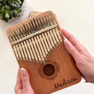 Puede incluir: Un kalimba de madera con 17 teclas, un diseño floral decorativo y el nombre "Madison" grabado en la parte delantera. El kalimba está hecho en China.