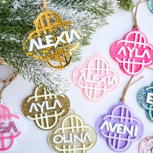 May include: A collection of personalized Christmas ornaments in various colors, including gold, pink, and purple. Each ornament is clover-shaped and features a name in white lettering. They hang from twine strings against a backdrop of a snowy pine branch.