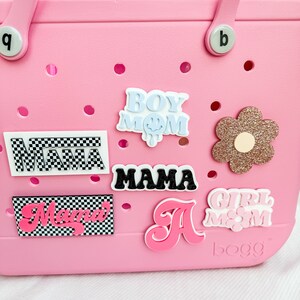 Pu&ograve; includere: Borsa tote rosa con varie toppe colorate, tra cui "Boy Mom", "Mama", "Girl Mom" e una lettera rosa "A". La borsa ha un motivo a scacchi bianco e nero sul fondo e la parola "BOGG" &egrave; stampata in basso a destra.