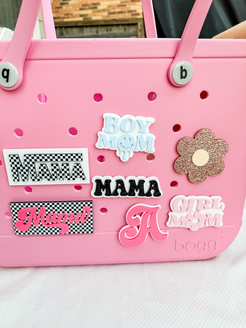 Pu&ograve; includere: Borsa tote rosa con varie toppe colorate, tra cui "Boy Mom", "Mama", "Girl Mom" e una lettera rosa "A". La borsa ha un motivo a scacchi bianco e nero sul fondo e la parola "BOGG" &egrave; stampata in basso a destra.