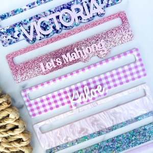 May include: A set of rectangular acrylic mahjong tile racks featuring various designs including blue and purple glitter, pink glitter with the text Let's Mahjong, purple and white gingham with the name Khloe, and white marble patterns with purple accents.