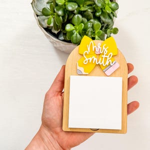 Könnte beinhalten: Ein Holz-Schreibtischaccessoire mit gelber Schleife und Bleistift-Design, mit dem Namen "Mrs. Smith" in weißer Schrift. Es enthält einen weißen Notizblock und wird in einer Hand gehalten. Im Hintergrund befindet sich eine Sukkulentenpflanze.