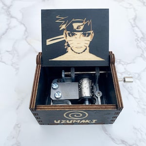 May include: A wooden music box with a hinged lid featuring a laser-etched image of an anime character. The box is dark brown with the word "UZUMAKI" and a spiral design on the front. The interior mechanism is visible.