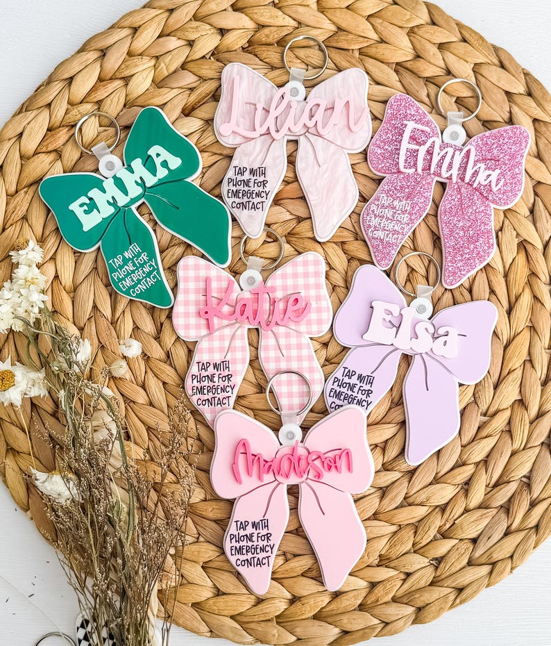 May include: A selection of bow-shaped keyrings in shades of green, pink, and lilac. Each keyring is personalized with a name and the text "TAP WITH PHONE FOR EMERGENCY CONTACT". The keyrings are displayed on a woven, circular mat.