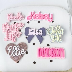 Può includere: Una collezione di targhette con nome in acrilico personalizzate e spille decorative con vari design, tra cui un cartello Nurse Life a quadretti rosa, un fiocco viola con il nome Elsa, un pezzo glitterato John 3:16 e un cuore glitterato oro rosa con Ellie.