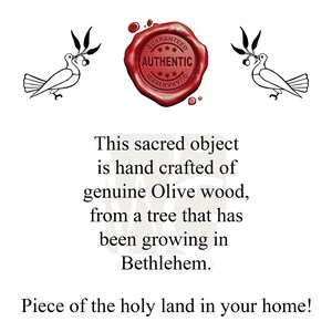 May include: A red wax seal with the words "GUARANTEED***AUTHENTIC" around a five-pointed star. Two white doves with olive branches in their beaks are on either side of the seal. The text below the seal reads: "This sacred object is hand crafted of genuine Olive wood, from a tree that has been growing in Bethlehem. Piece of the holy land in your home!"