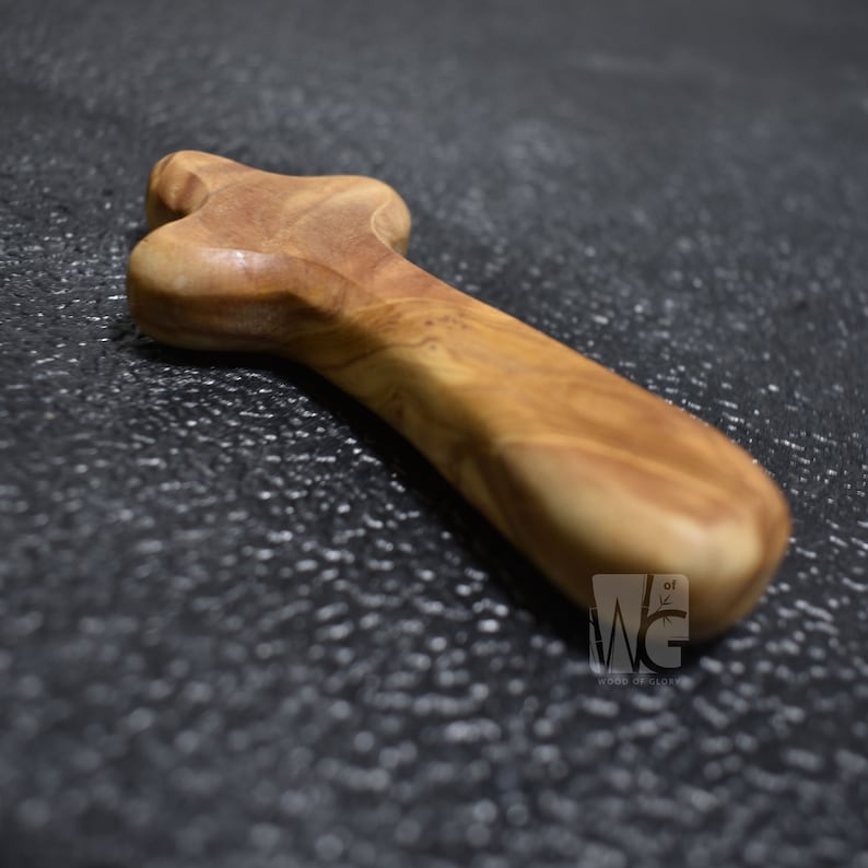 May include: A wooden prayer cross made of olive wood. The cross is a simple, traditional design with a rounded top and a long, slender handle. The cross is light brown in color and has a smooth, polished finish.