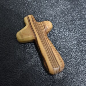 May include: A wooden cross made of olive wood with a natural wood grain pattern. The cross is a simple, traditional design with a rounded top and bottom.