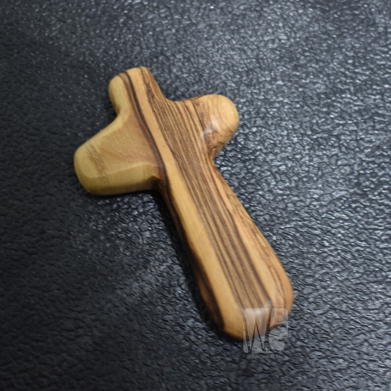 May include: A wooden cross made of olive wood with a natural wood grain pattern. The cross is a simple, traditional design with a rounded top and bottom.
