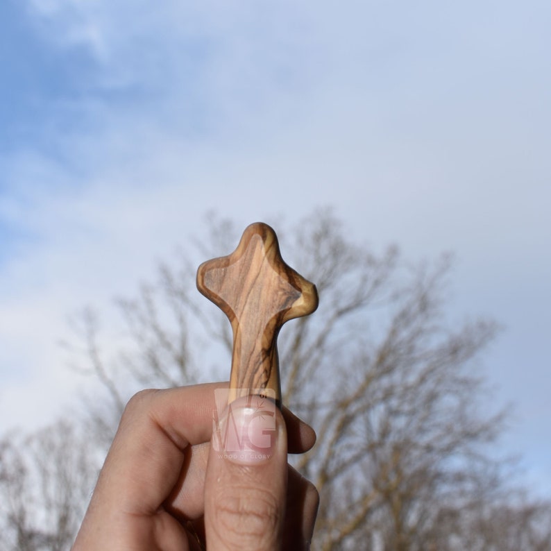 May include: A hand holds a small wooden cross with a light brown and dark brown wood grain pattern. The cross is against a blue sky with white clouds and a blurred background of bare tree branches.