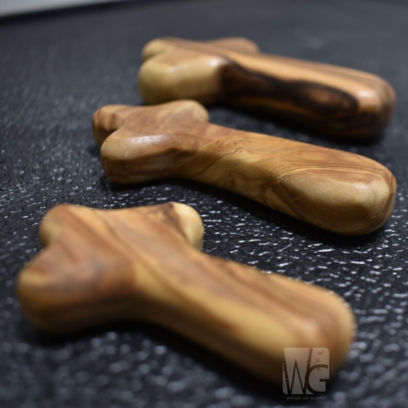 May include: Three wooden olive wood crosses, each with a unique grain pattern. The crosses are arranged in a row, with the largest cross in the back and the smallest cross in the front. The crosses are made of a light brown wood with darker brown streaks.