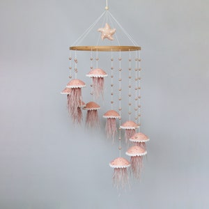 May include: A pink and white baby mobile with a wooden ring and a starfish hanging from the top. The mobile features eight pink jellyfish with white dots and pink and white beads hanging from the ring.