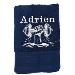 Può includere: Asciugamano blu navy con il nome "Adrien" in lettere bianche sopra una grafica bianca del braccio di un sollevatore di pesi che tiene un bilanciere. L'asciugamano è piegato e ha un aspetto strutturato.