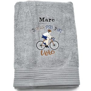 Op de afbeelding: Grijze badhanddoek met geborduurde tekst en een fietserontwerp. De tekst "Marc" staat boven de zin "Je peux pas j'ai vélo" en een fietser op een fiets. De handdoek heeft een geribbelde rand.