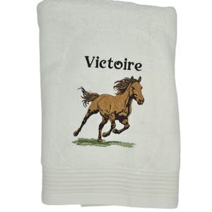 Peut inclure: Serviette de bain blanche avec un cheval brun brodé courant et le nom "Victoire" en lettres noires.