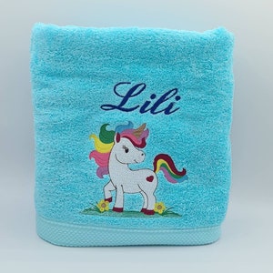 Peut inclure: Une serviette de bain bleu clair avec une licorne blanche brodée dessus. La licorne a une crinière et une queue arc-en-ciel. La serviette porte également le nom "Lili" brodé.