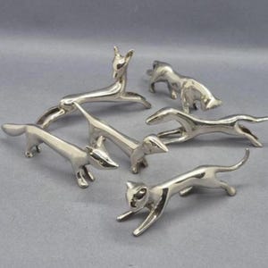 May include: A collection of six polished silver-toned metal animal figurines, including a deer, a cat, and several dog-like shapes, each depicted in a dynamic, elongated pose with smooth, reflective surfaces and minimalist, stylized features.