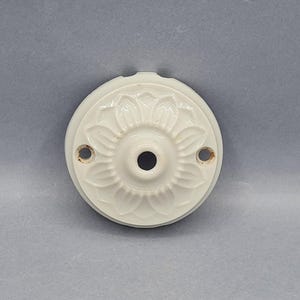 May include: Cream-coloured ceramic ceiling rose with a floral pattern. The circular plate features a central hole and two smaller mounting holes. The raised floral design is detailed, and the fixture has a vintage appearance.
