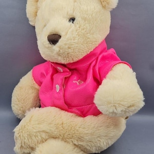 May include: A plush teddy bear with cream-colored fur and a brown nose. The bear is wearing a bright pink jacket with white buttons. The toy is sitting against a gray background.