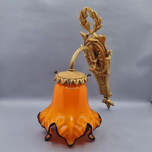 Könnte beinhalten: Antike Wandleuchte mit orangefarbenem Glasschirm und goldfarbenem Metallrahmen. Der Schirm hat ein gerüschtes Design mit schwarzem Besatz. Die Leuchte hat eine dekorative Rückplatte und einen gebogenen Arm mit Lorbeerkranz-Detail.