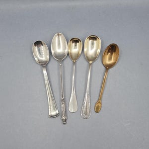 May include: Five antique spoons of varying designs and sizes are arranged against a gray background. The spoons are silver and gold-toned, showcasing different handle and bowl shapes. The spoons are likely for serving or eating.
