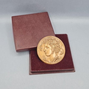 May include: A bronze-colored round medallion featuring a classical profile, resting in a burgundy presentation box. The medallion has intricate details and is displayed on a burgundy velvet-lined interior.