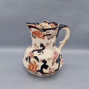 Puede incluir: Una jarra de cerámica blanca con un patrón floral azul y dorado. La jarra tiene una boca ancha y un asa.