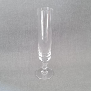 May include: A clear champagne flute with a tall, cylindrical bowl. The glass has a round base and a decorative stem. The background is a solid grey.