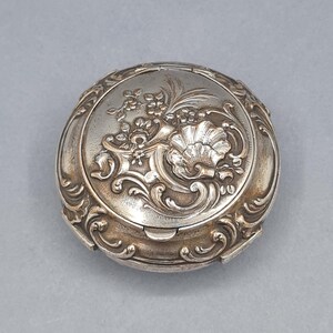Puede incluir: Una caja redonda antigua de color plateado con intrincados detalles en relieve floral y con forma de concha. La tapa presenta un diseño central de concha rodeado de hojas y flores. La caja tiene una tapa con bisagras y un borde decorativo.