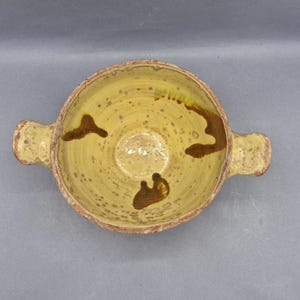 Może przedstawiać: Okrągła ceramiczna miska z dwoma bocznymi uchwytami, posiadająca żółte szkliwione wnętrze z brązowymi nakrapianymi wzorami oraz trzema wyraźnymi ciemnobrązowymi abstrakcyjnymi oznaczeniami. Zewnętrzna krawędź i uchwyty mają teksturowane, ziemiste wykończenie.