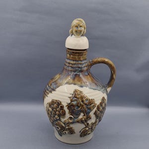 Peut inclure: Une carafe à vin en céramique avec un glaçage brun et bleu. La carafe a une poignée et un motif de vigne sculpté. Le bouchon présente une petite figurine en céramique d'une femme aux cheveux blonds.
