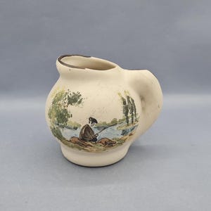 Puede incluir: Jarra de cerámica color crema con cuerpo redondeado y asa. La jarra presenta una escena pintada a mano de una persona pescando junto a un río, con árboles y agua de fondo. El borde está rematado con una línea marrón.