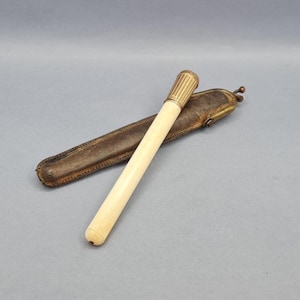 May include: An antique ivory-colored writing instrument with a gold-toned cap, resting on a brown leather case. The pen has a cylindrical shape with a ribbed cap design. The leather case is rectangular with a rounded end.