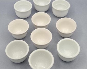 May include: A collection of nine small, white ceramic bowls arranged in a grid pattern. The bowls have a rounded shape with a smooth, glossy finish. They are likely used for serving small portions of food or as decorative items.