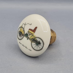 Könnte beinhalten: Cremefarbener Keramik-Schrankknopf mit einem Vintage-Fahrrad-Design mit roten und gelben Akzenten. Der Knopf hat einen Messingfuß und den Text "Ford's First Car, 1896".