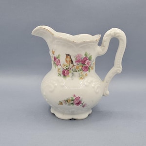 Puede incluir: Una jarra de crema de porcelana blanca con un diseño floral y un pequeño pájaro posado en una rama. La jarra tiene un asa delicada y un cuerpo redondeado.