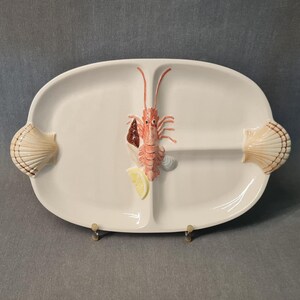 Peut inclure: Plat à servir en céramique blanche divisé en deux sections. Il présente un homard et une tranche de citron en relief, ainsi que des poignées en forme de coquillage. Le plat mesure environ 36 cm de long et 23 cm de large.