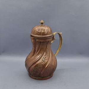 May include: A copper jug with a brass handle and lid. The jug features an ornate design with floral motifs and a swirling pattern.