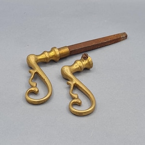 Può includere: Due antiche maniglie per cassetti in ottone con un design a volute decorativo. Una maniglia è attaccata a un manico in legno marrone. L'altra maniglia è separata. L'hardware è di un caldo colore oro.