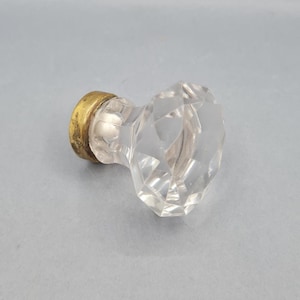 May include: A clear, faceted glass door knob with a brass base. The knob has a diamond shape and is designed for a door. The brass base shows signs of age and wear.