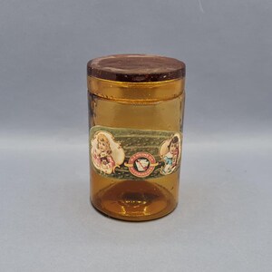 Op de afbeelding: Een vintage amber glazen pot met een houten deksel en een etiket met twee kinderen. Het etiket vermeldt "The Mikine Co. Philadelphia".