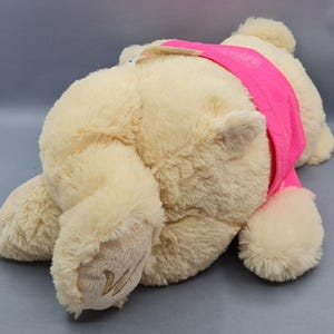 May include: A plush, cream-colored teddy bear wearing a bright pink band across its chest. The bear is lying down, with its paws and head visible. The fur is soft and fluffy, and the bear appears to be new.