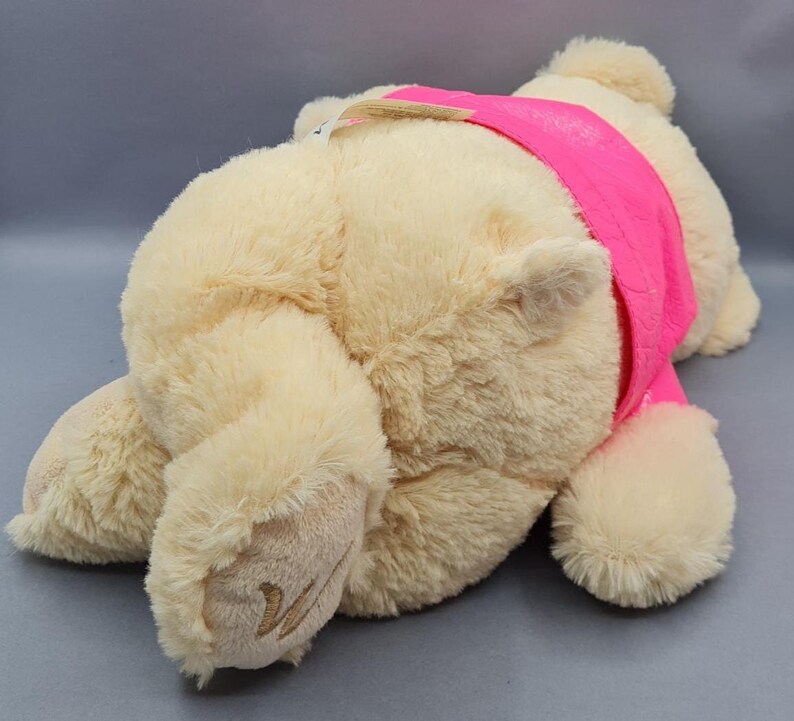 May include: A plush, cream-colored teddy bear wearing a bright pink band across its chest. The bear is lying down, with its paws and head visible. The fur is soft and fluffy, and the bear appears to be new.