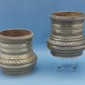 Puede incluir: Dos jarrones antiguos de metal plateado con intrincados diseños en relieve. Los jarrones tienen forma cilíndrica con una base más ancha y una abertura más estrecha. Los diseños incluyen patrones geométricos y motivos florales. Los jarrones se exhiben sobre una superficie transparente sobre un fondo azul.