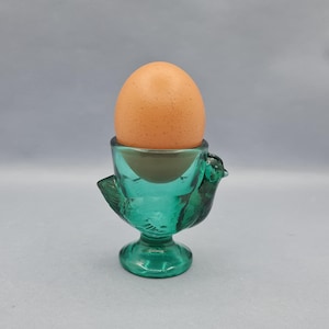 Puede incluir: Un huevero de vidrio de color turquesa con forma de pájaro, que sostiene un huevo marrón. El huevero tiene un diseño detallado de pájaro con alas y una base de pedestal. El huevo es de color marrón claro.
