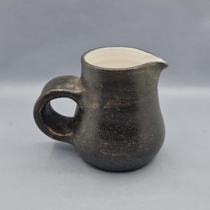 Puede incluir: Un pequeño jarro de cerámica hecho a mano con exterior marrón oscuro e interior color crema. El jarro tiene un cuerpo redondeado, un asa curva y un pico para verter. La superficie tiene un aspecto texturizado.